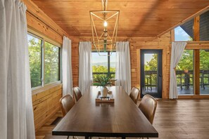 Ein Holzhütten-Interieur mit Esstisch, Stühlen, einer Hängeleuchte und großen Fenstern mit Vorhängen.