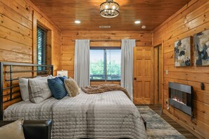 Ein gemütliches Schlafzimmer in einer Holzhütte mit Bett, Kamin und großem Fenster mit Vorhängen.