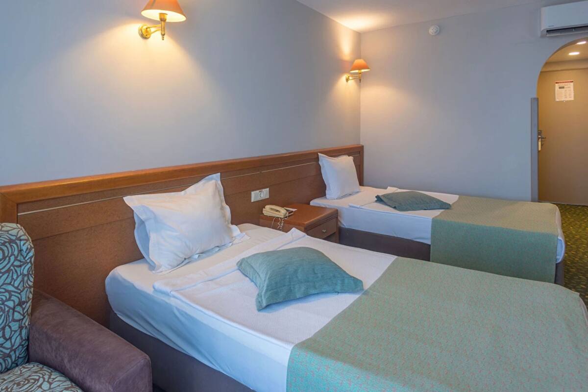 A hotel room with two single beds, each with a pillow and a blanket. There is a telephone on the nightstand, a chair with a patterned cushion, and a door with a sign.