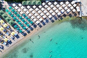 Vista aérea de una playa con hileras de sombrillas blancas y techos verdes, agua turquesa cristalina y gente nadando.