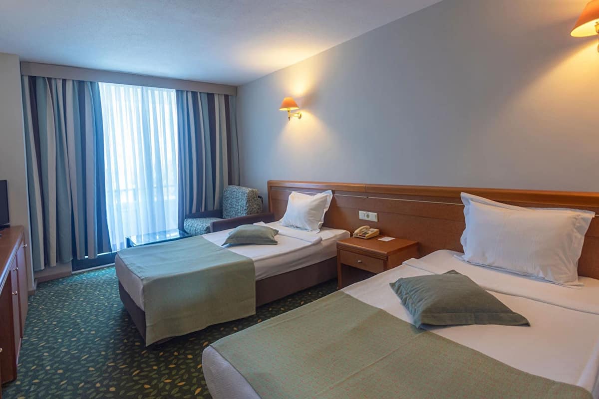 A hotel room with two beds, a desk, a chair, a lamp, and a window with curtains.