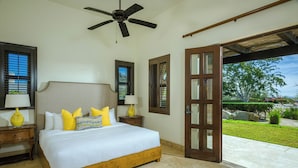 Une chambre à coucher avec un lit, une tête de lit en bois, deux tables de chevet avec des lampes, un ventilateur de plafond et une vue sur l’extérieur par une porte.