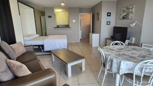 Un salon moderne avec un canapé en cuir marron, une petite table basse en bois, une table à manger avec des chaises blanches, un téléviseur à écran plat et un grand miroir.
