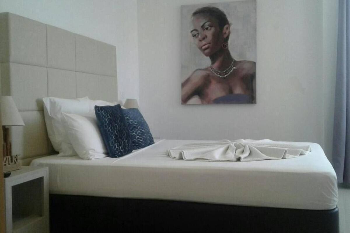Una cama bien hecha con almohadas blancas y azules, una toalla doblada y un cuadro de una mujer en la pared.