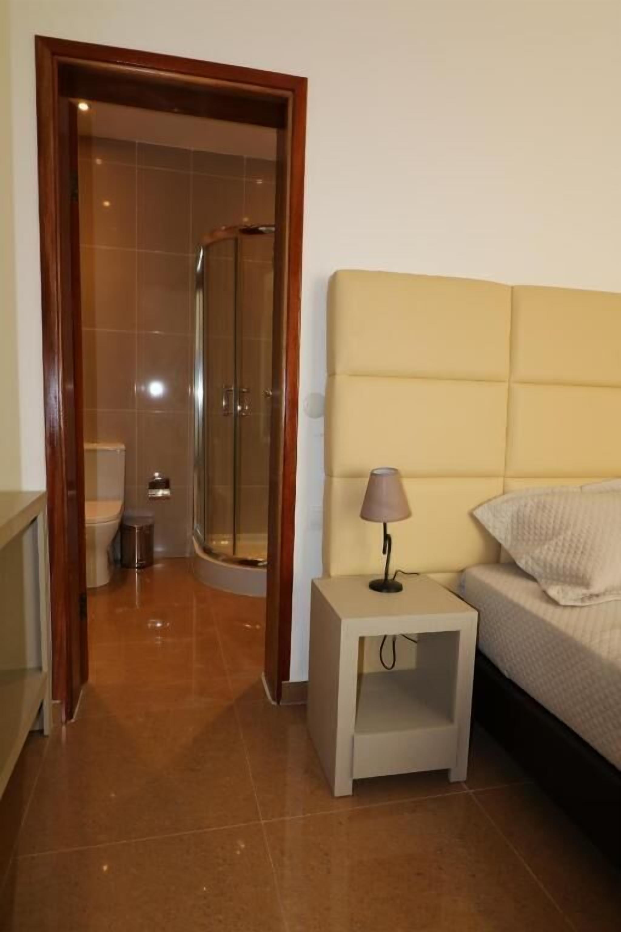 A bedroom with a bed, a nightstand with a lamp, a wooden door leading to a bathroom with a shower, and a tiled floor.