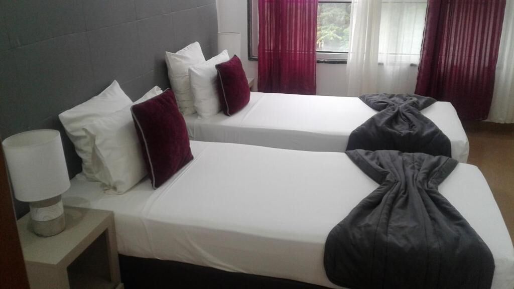 Een hotelkamer met twee bedden, elk voorzien van witte lakens en een donkerde plaid. Er staat een nachtkastje met een lamp en er is een raam met rode gordijnen.
