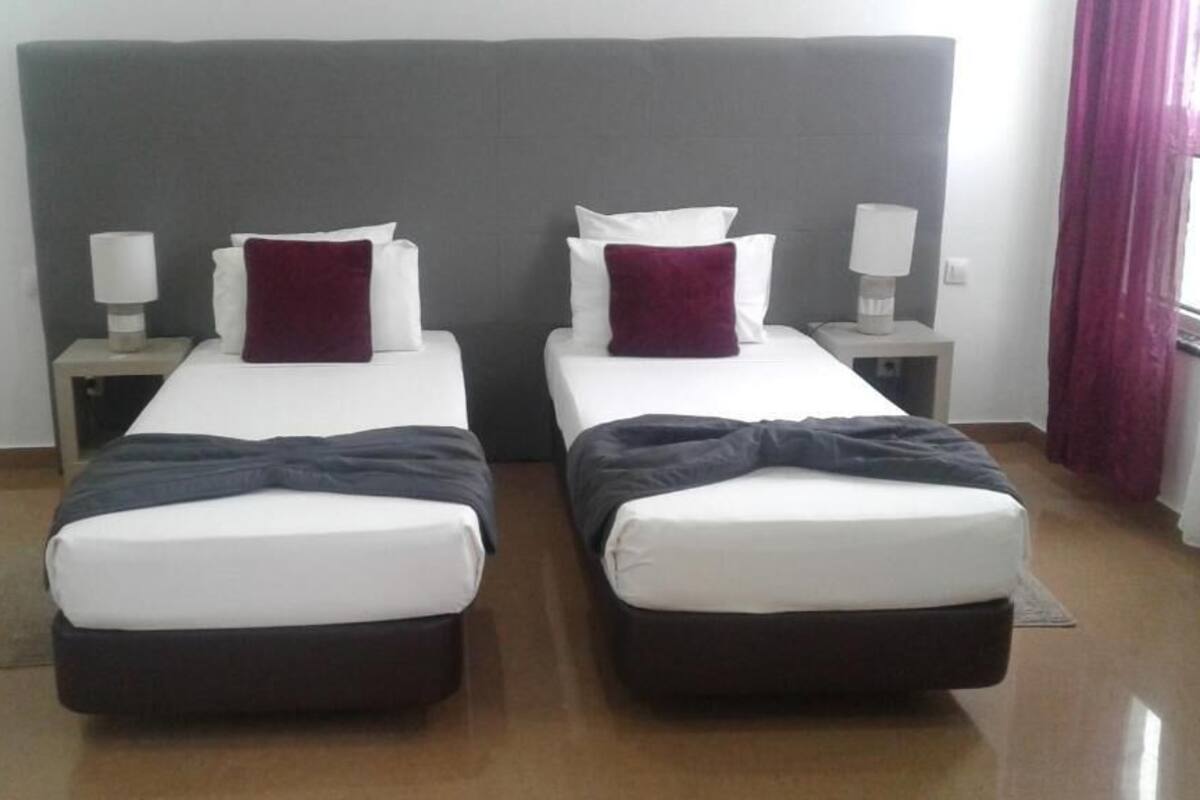 Habitación doble con dos camas individuales, cada una con colchón blanco y cabecero oscuro. Hay dos mesitas de noche con lámparas y una ventana con cortinas.