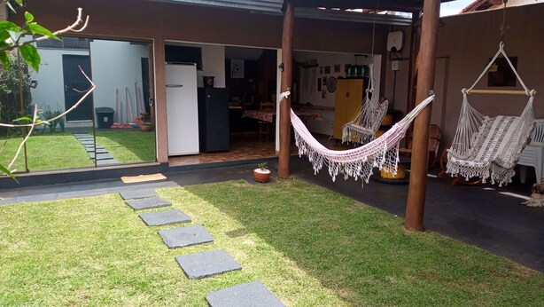 Ein überdachter Außenbereich mit einer Hängematte, ein kleiner Garten mit trittfesten Steinen und Blick in ein Haus mit Küche und Essbereich.