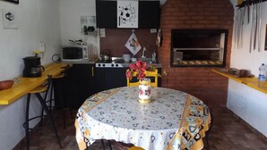 Eine kompakte Küche mit Mikrowelle, Gasherd und einem Backofen aus Ziegelsteinen. Ein Esstisch mit gemustertem Tischläufer und eine Blumenvase sind ebenfalls vorhanden.