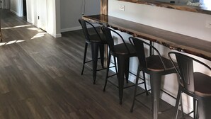 Une cuisine moderne avec un comptoir-bar en bois, quatre tabourets de bar, un téléviseur à écran plat fixé au mur et une grande horloge.