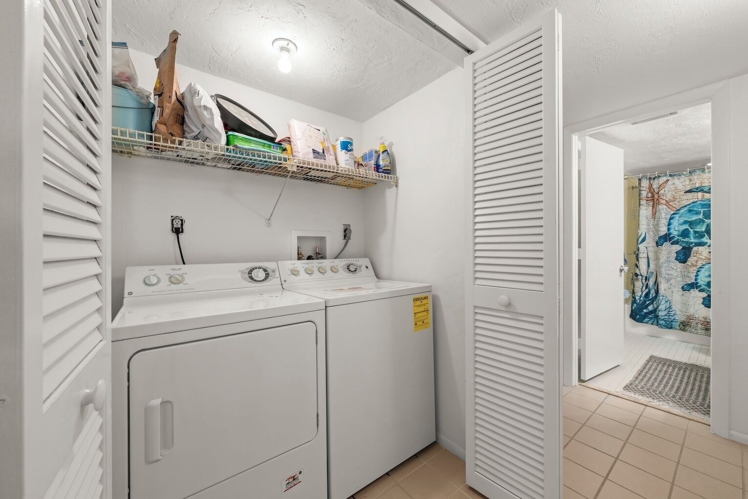 Lavandería con dos lavadoras y secadoras blancas, un estante superior con diversos objetos, y una puerta que conduce a otra habitación con una cortina estampada.