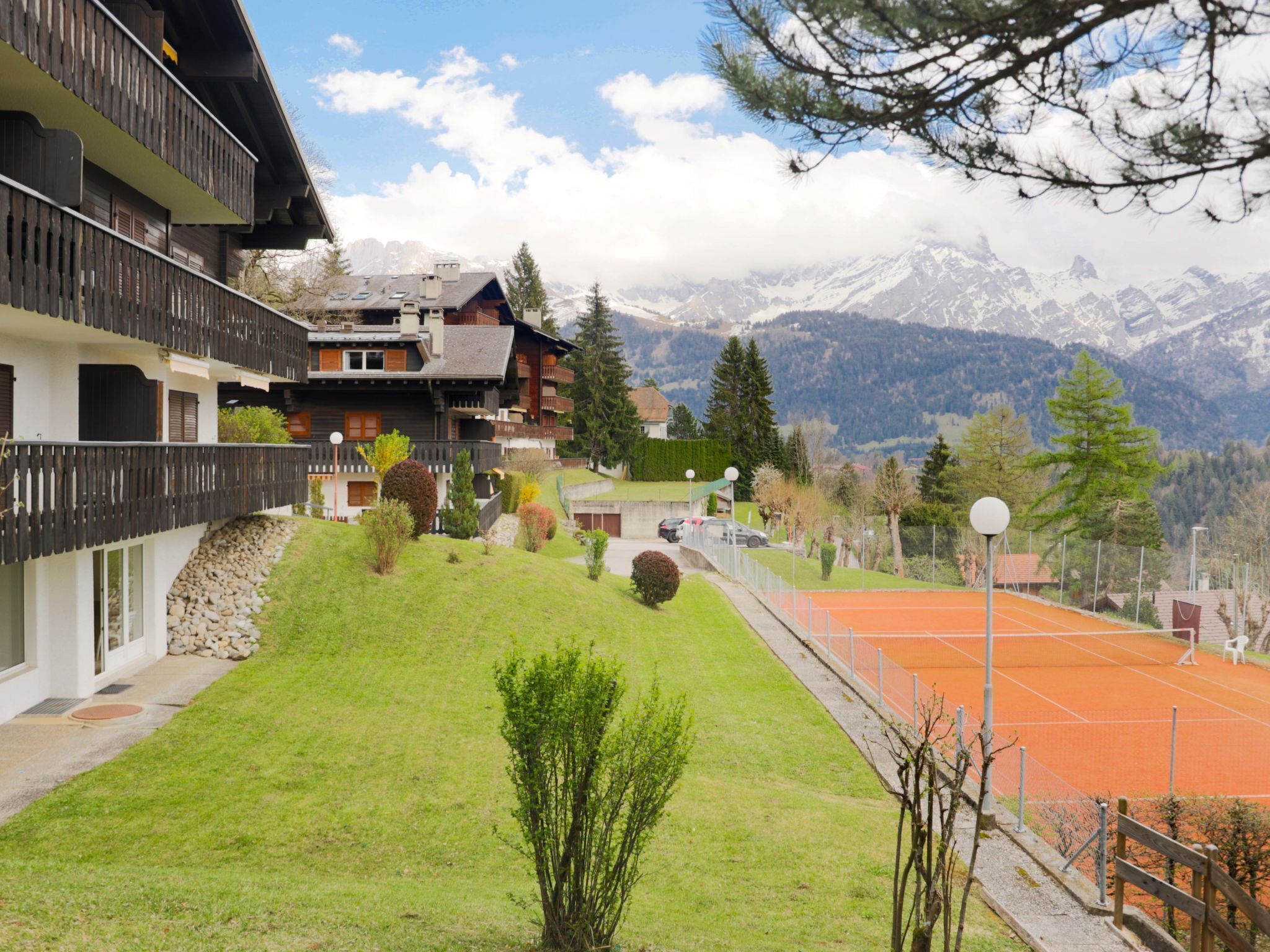 A chalet-style building with a balcony, a tennis court, and a mountainous backdrop.