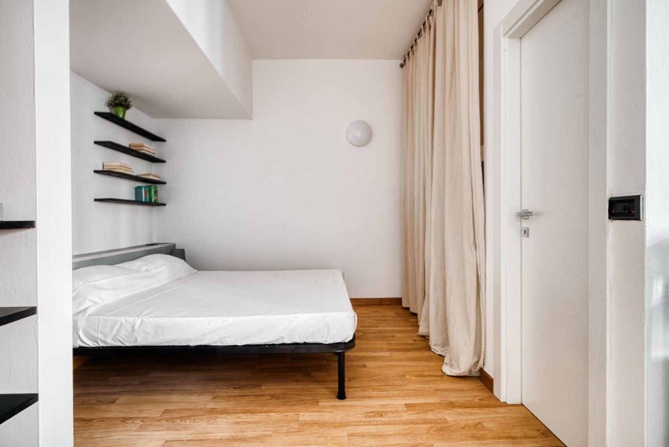 A bedroom with a bed, wooden flooring, a white wall, a ceiling light, and a door with curtains.