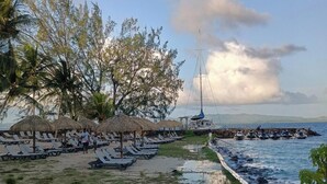 Ein Strand mit Reetdachschirmen, Liegestühlen und einem Segelboot.