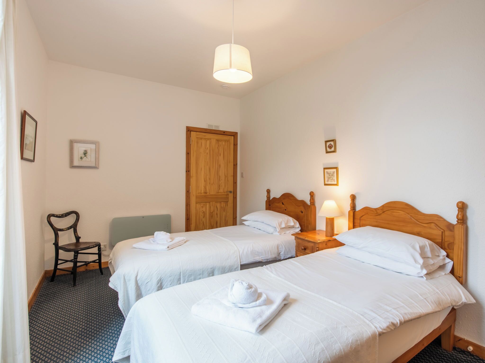 A hotel room with two single beds, a wooden wardrobe, a chair, and a small table with a lamp.