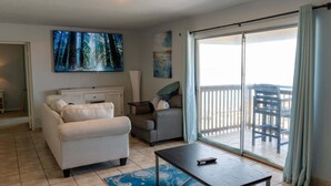 Ein Wohnzimmer mit einer Couch, einem Couchtisch und einem Fernseher. Eine Glastür führt auf einen Balkon mit Gartenmöbeln.