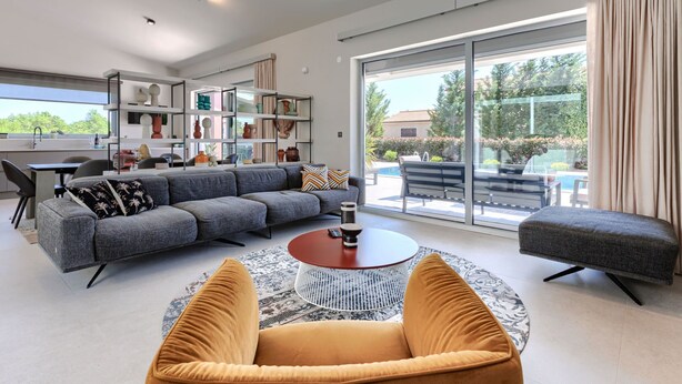 Una sala de estar moderna con un sofá gris, una mesa de centro redonda y un ventanal con vista a una piscina.