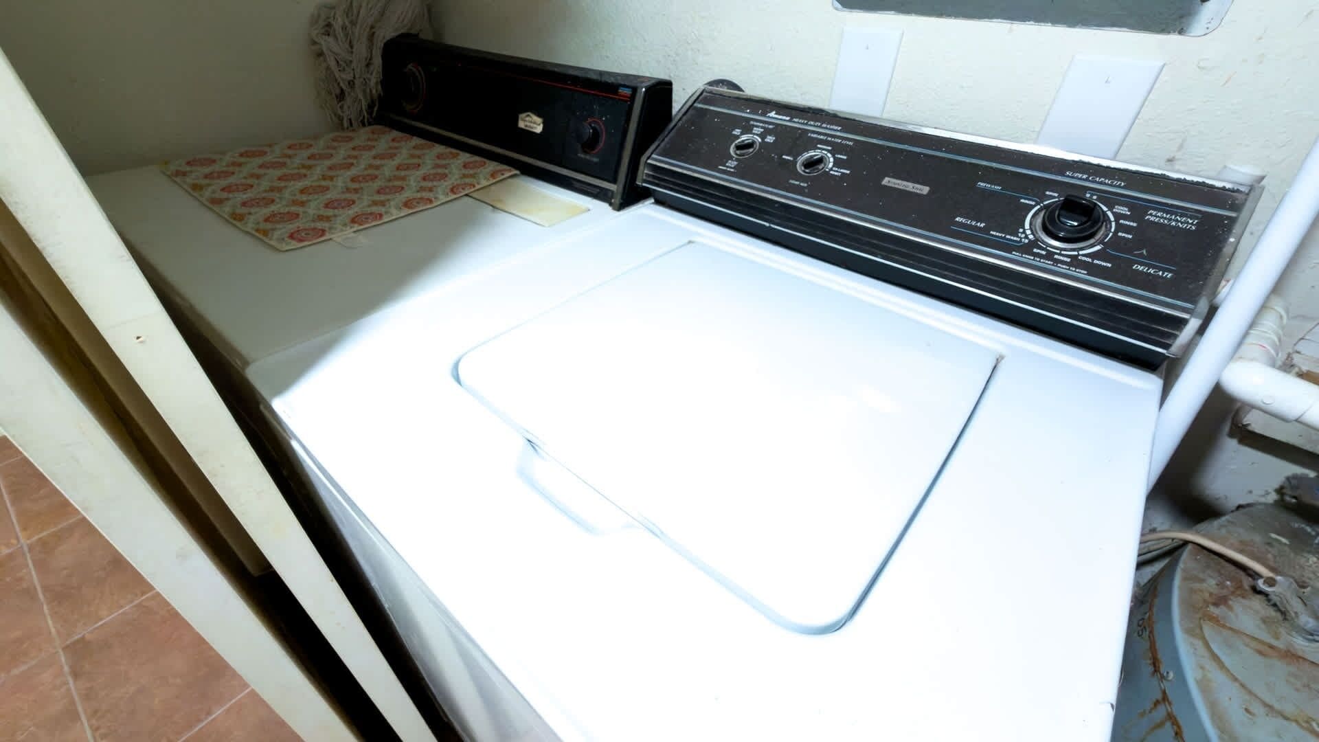 A white washing machine with a control panel, a patterned mat, and a tiled floor.