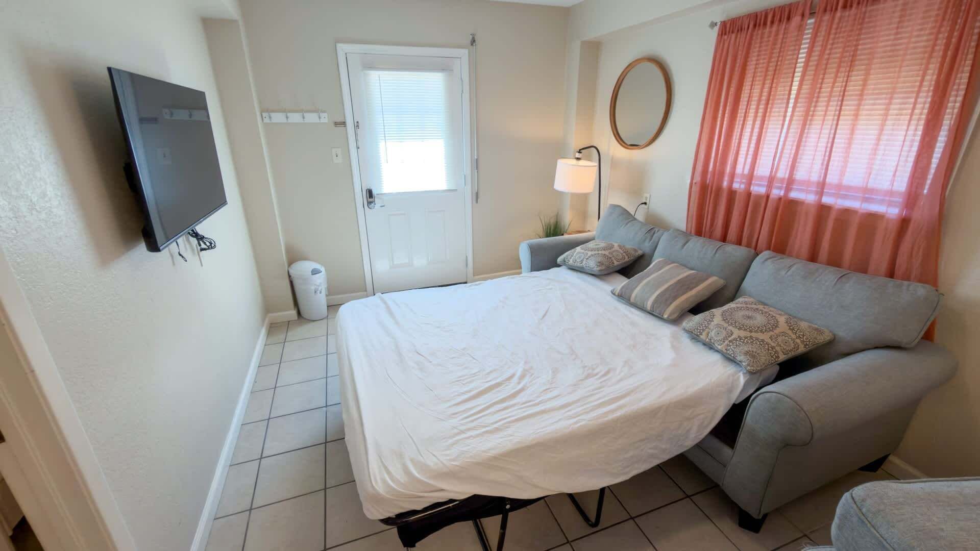 A bedroom with a bed, a grey sofa, a TV mounted on the wall, a door with a window, and a lamp.