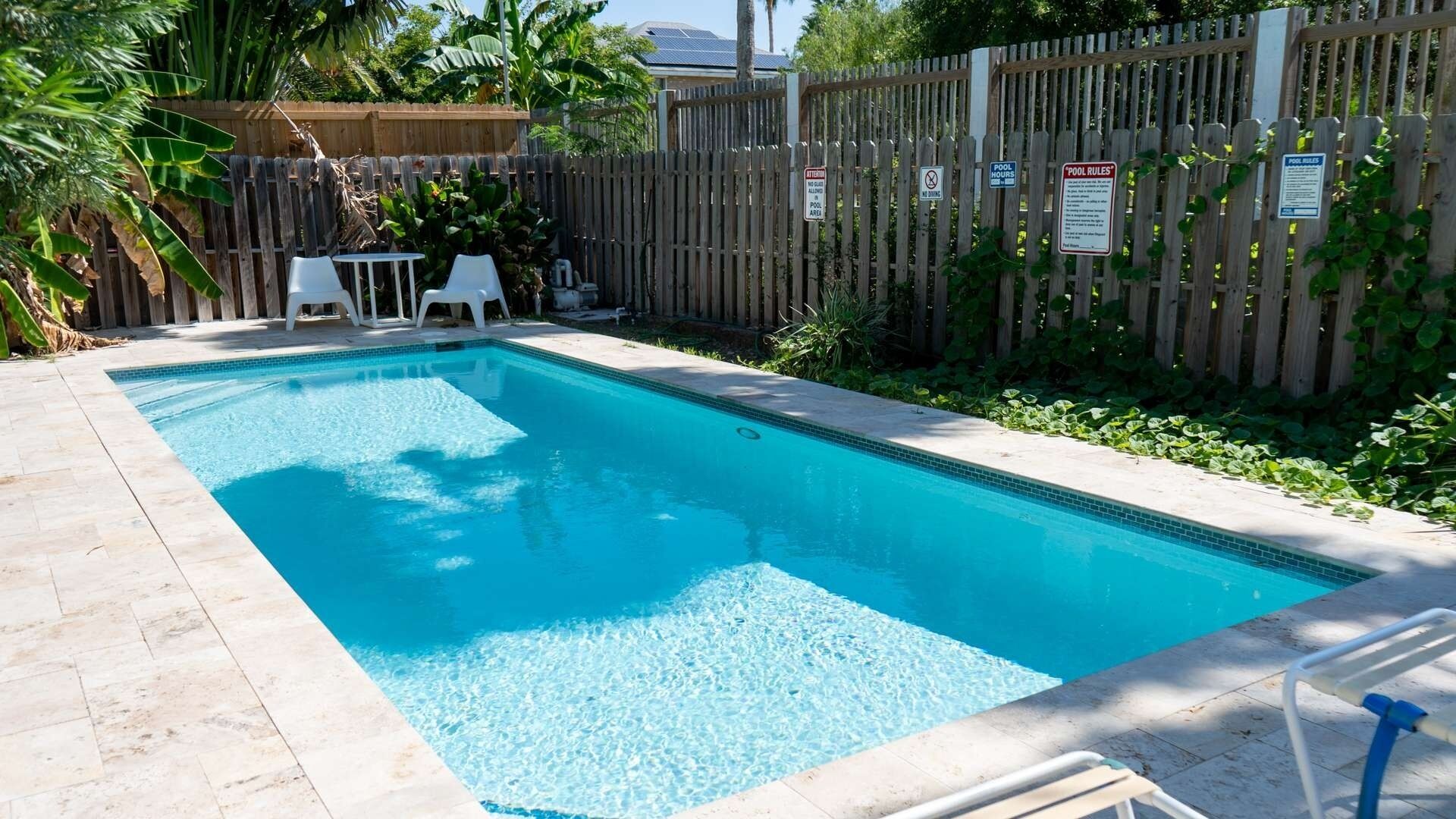 A rectangular swimming pool with a tiled edge, surrounded by a wooden fence, and a small patio area with a table and chairs.