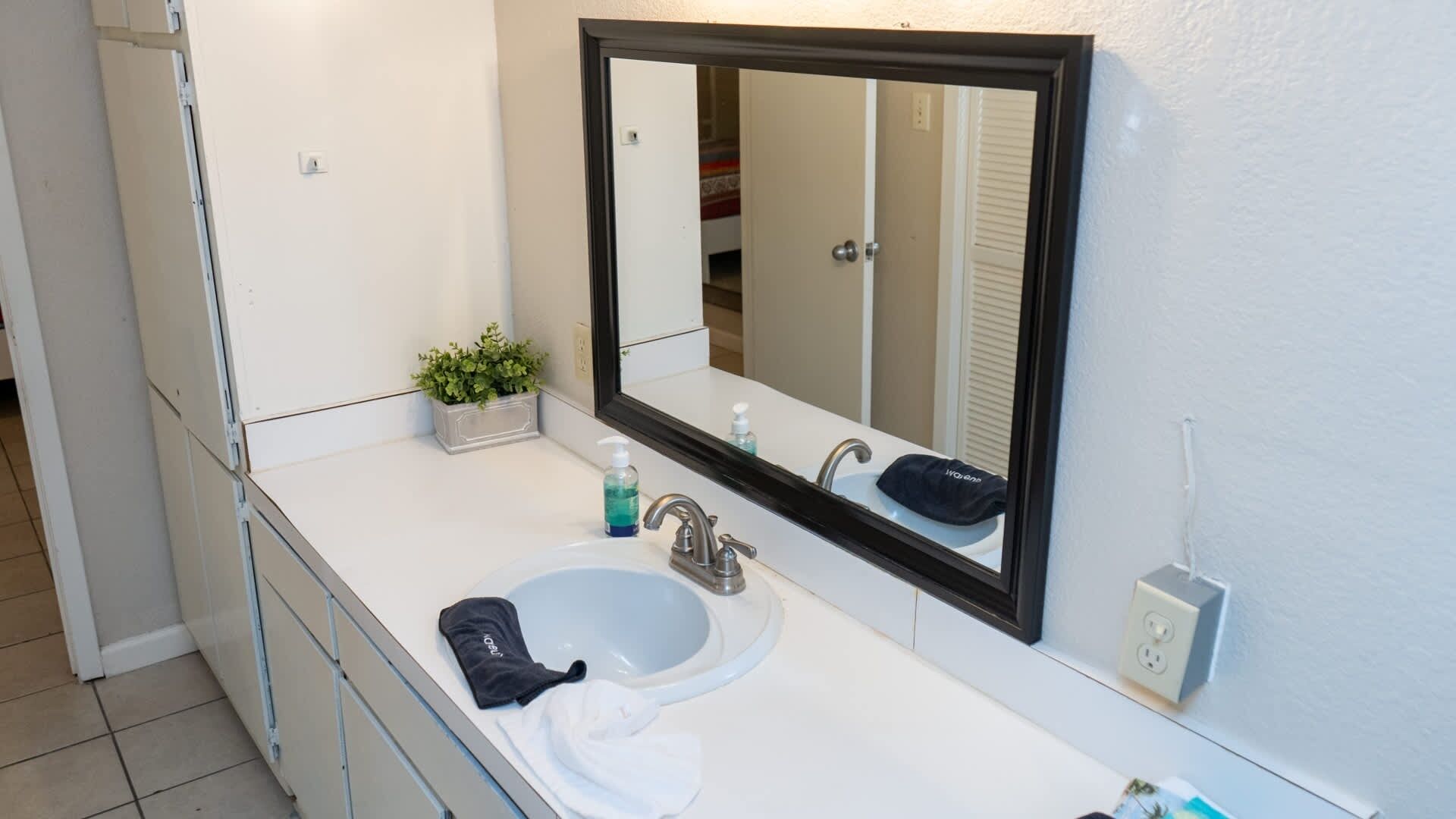 Ein Badezimmer mit einem weißen Waschbecken, einem spiegelnden Schwarzrahmen, einer kleinen Pflanze und einem Handtuch auf der Ablagefläche.