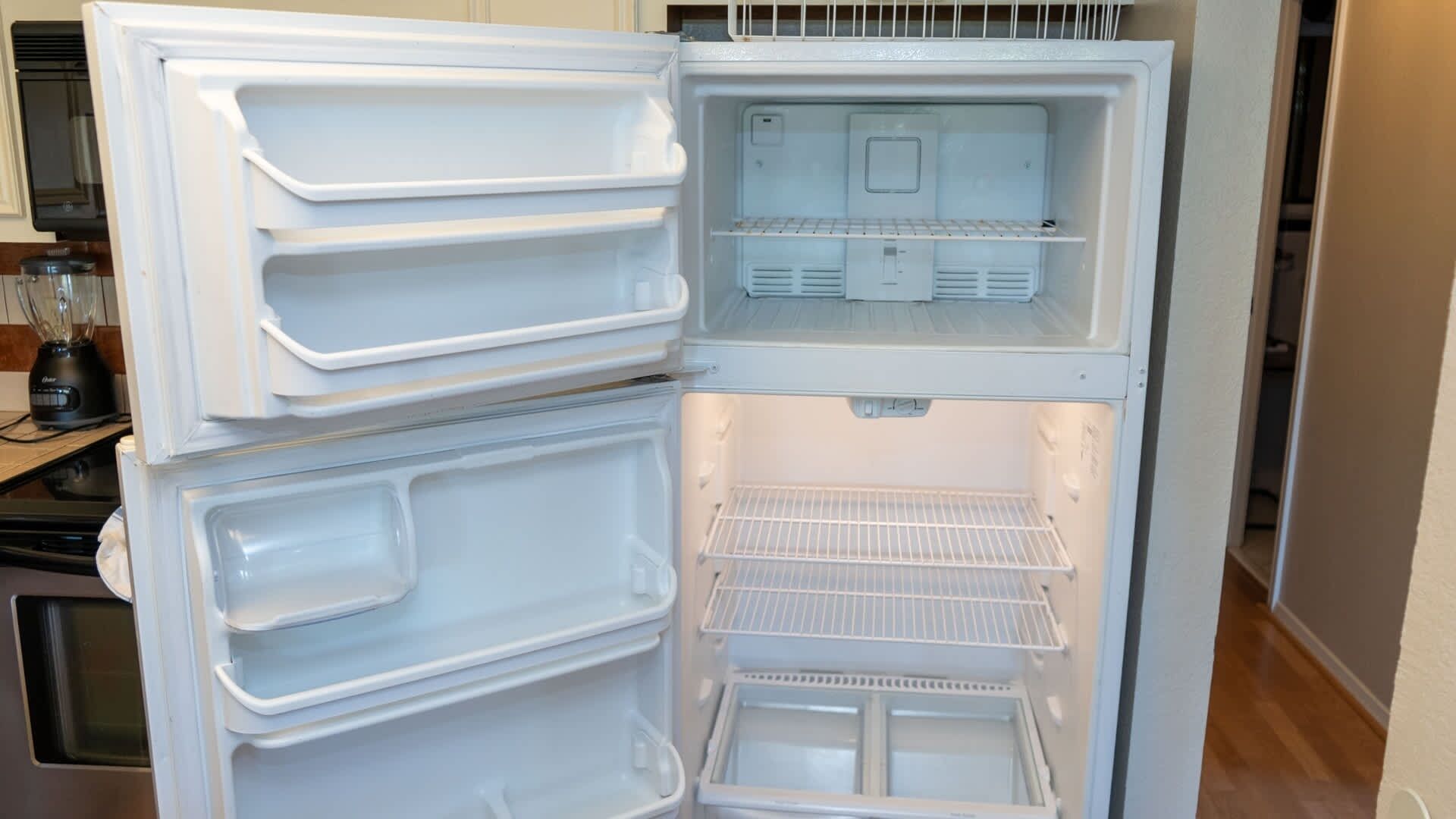 Avoin jääkaappi, jossa on useita hyllyjä ja lokeroita, ja joka sijaitsee keittiössä, jossa on näkyvillä myös tehosekoitin ja muita kodinkoneita.