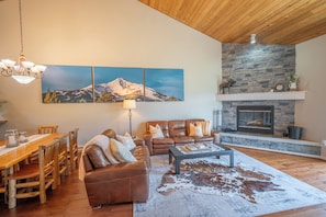 A living room with a fireplace, a dining area with a wooden table and chairs, and a large wall-mounted painting of a mountain. - Candlelight Meadows Escape | Hot Tub Near Town Center & Spacious Great Room (Big Sky)