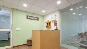 A reception area with a wooden desk, a reception sign, a small shelf with items, and a glass partition. - A Square Homes & Stay (New Delhi)
