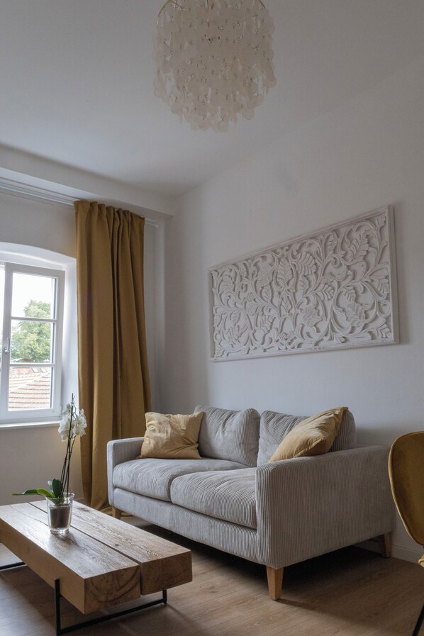 A living room with a sofa, a coffee table, a chair, a window with curtains, and a decorative wall piece. - ღLiboria | Casa Mangifera (Murnau am Staffelsee)
