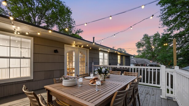 An outdoor deck with a wooden table and chairs, string lights, and a white fence. - The Blackmoore-Theater-GameRoom-Gym (Overland Park)