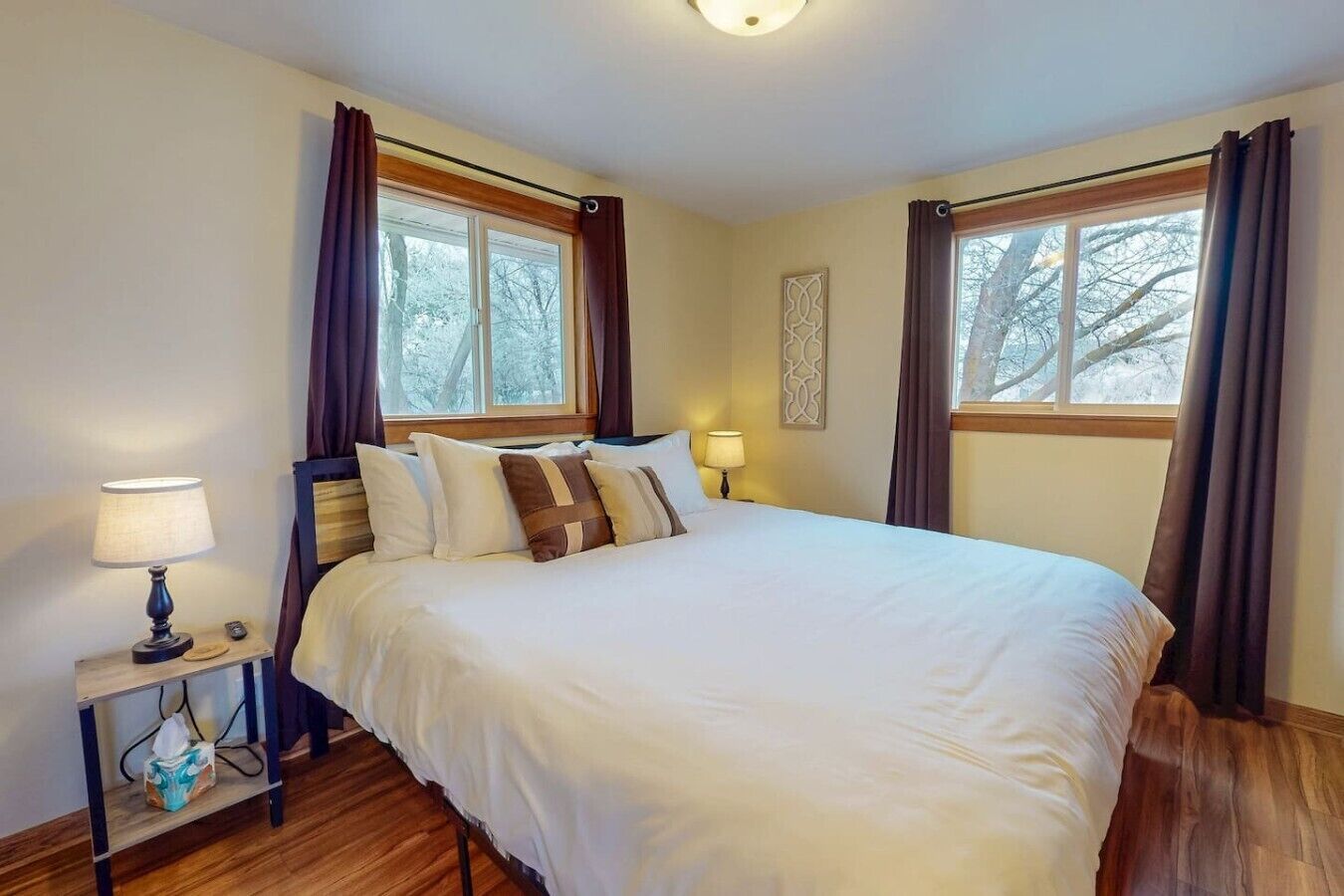 A neatly made bed with white bedding and brown pillows, two bedside lamps, a wooden nightstand with a tissue box, and a window with curtains.