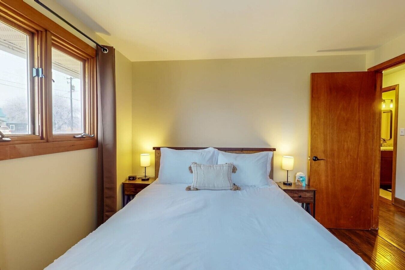 Une chambre d’hôtel avec un lit, deux tables de chevet avec des lampes, une porte en bois et une fenêtre avec des rideaux.