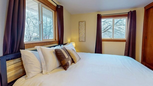 Une chambre d’hôtel avec un grand lit, deux fenêtres avec des rideaux, une lampe de chevet et un élément décoratif mural.