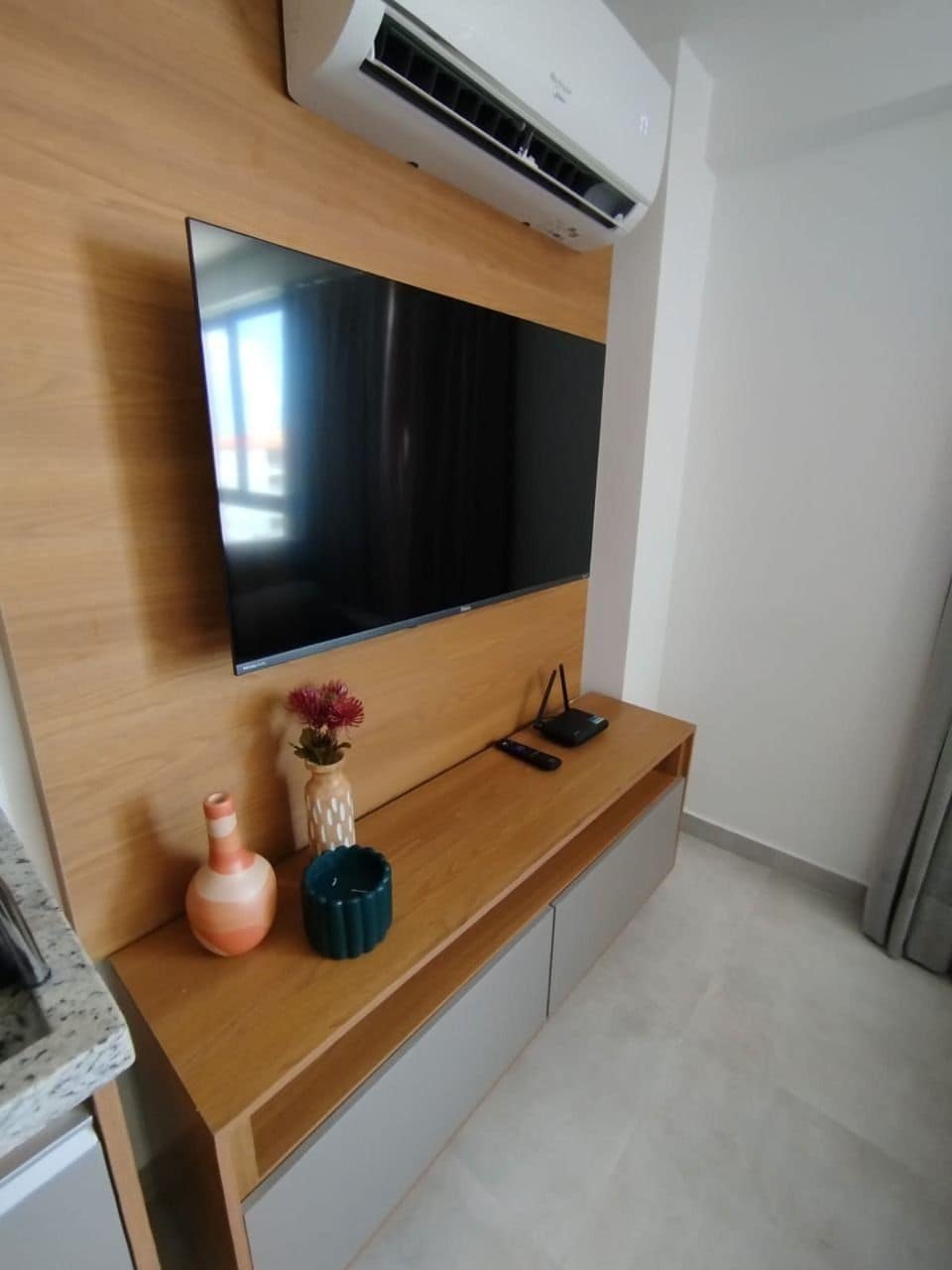 A modern living room with a flat-screen TV mounted on a wooden wall, a small table with decorative items, and a built-in air conditioning unit.