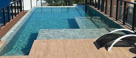 A rooftop infinity pool with wooden deck and lounge chairs, overlooking the ocean.