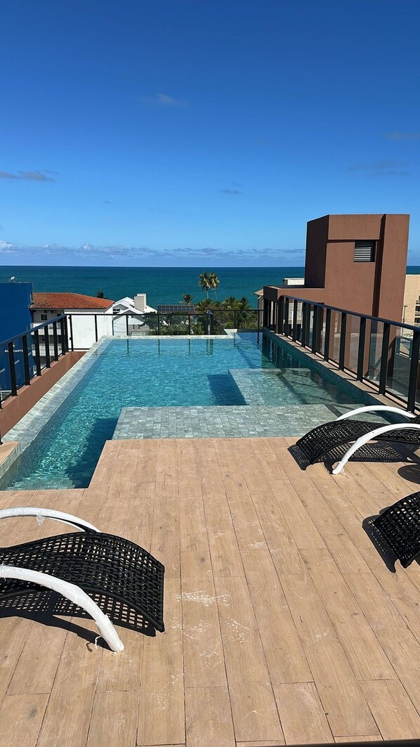 A rooftop infinity pool with wooden deck and lounge chairs, overlooking the ocean. - Carneiros Beach Exclusive (Tamandaré)