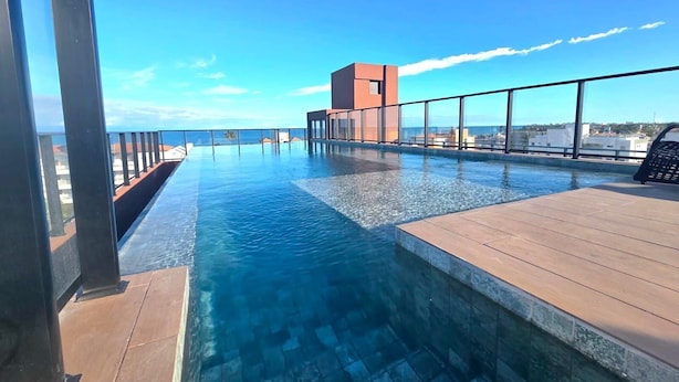 A rooftop infinity pool with a clear view of the ocean, a modern building, and a wooden deck. - Carneiros Beach Exclusive (Tamandaré)