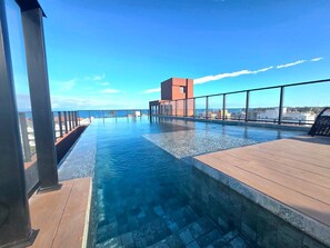 Ein Rooftop-Pool mit Infinity-Kante und freiem Blick auf den Ozean, ein modernes Gebäude und eine Holzterrasse.