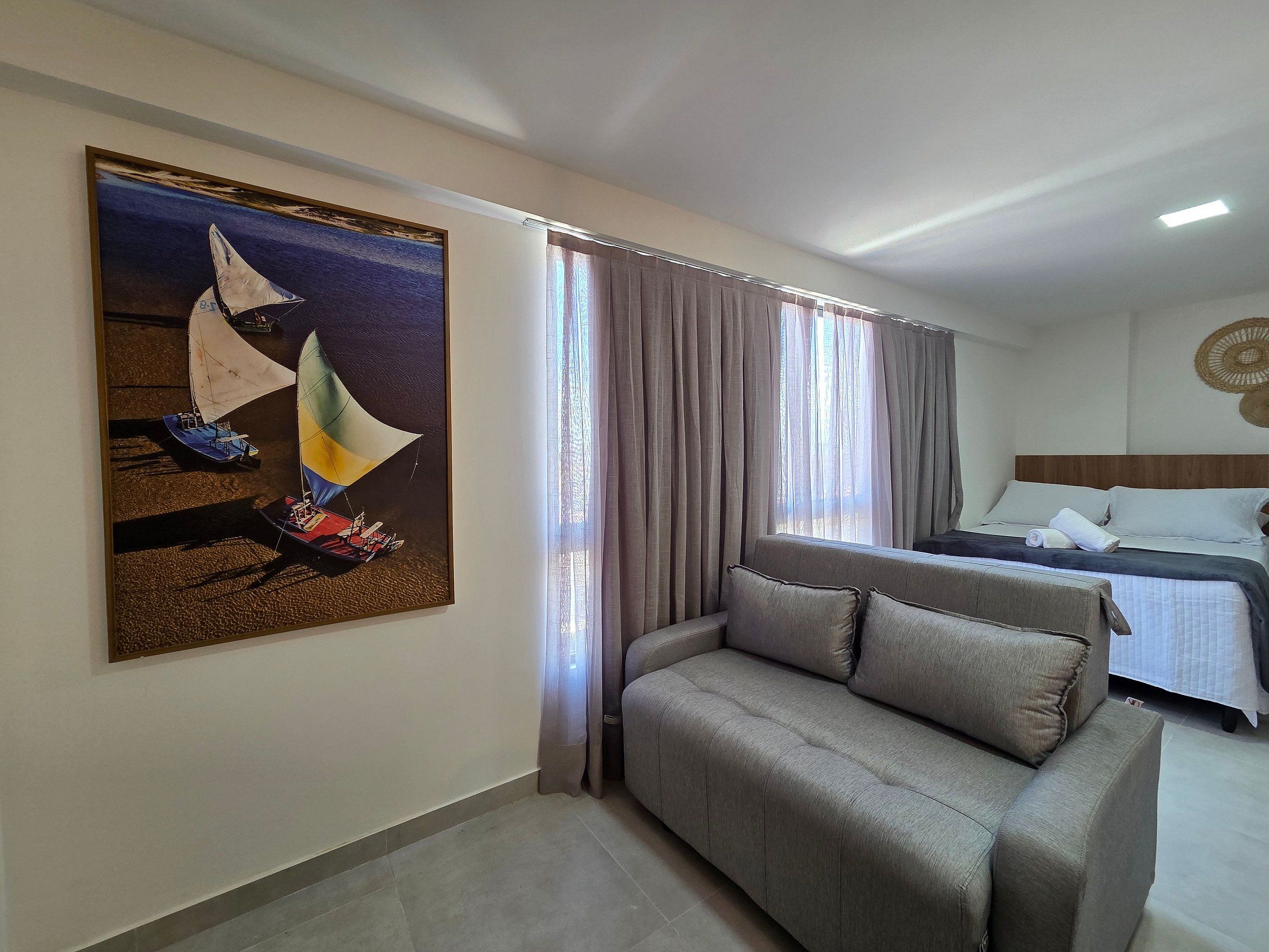 A hotel room with a bed, a sofa, a painting of a sailboat, and curtains.