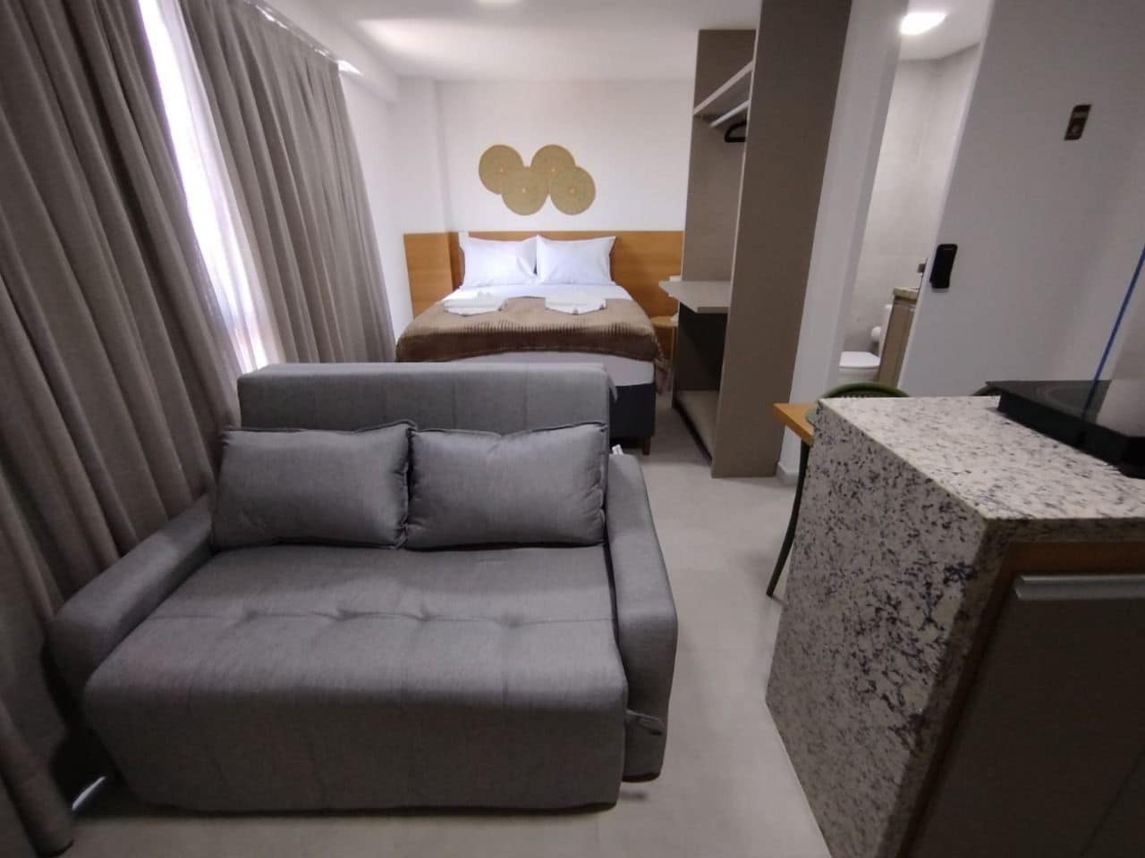 A modern hotel room with a grey sofa, a bed with a wooden headboard, a marble countertop, and a wall-mounted light fixture.