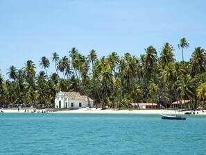 A beach with palm trees, a small white building, and a boat in the water. - Carneiros Beach Exclusive (Tamandaré)