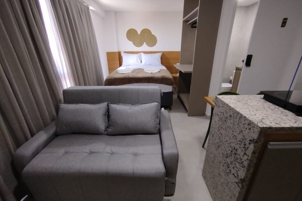 A modern hotel room with a grey sofa, a bed with a wooden headboard, a marble countertop, and a wall-mounted light fixture.