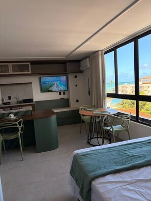 A hotel room with a bed, a dining area with chairs, a TV on the wall, and a view of the ocean. - Carneiros Beach Exclusive (Tamandaré)