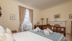 A hotel room with a large bed, a wooden headboard, a desk, a chair, a fireplace, and a window with curtains. - South Street (No 16A) | Elie (Elie)