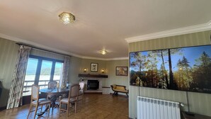 A living room with a fireplace, a dining table with chairs, a large TV screen, and a view of trees through the window. - Alma CaloRosa – Sea View Escape in Romantic Moledo, Northern Portugal (Moledo)