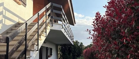 Un edificio moderno con una escalera que lleva a un balcón, rodeado de vegetación exuberante y follaje rojo.