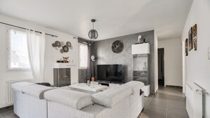 A modern living room with a grey sofa, a flat-screen TV, and a dining table. - New and cozy house for 6 with garden (Beaussais-Sur-Mer)