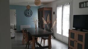 A dining area with a wooden table and chairs, a clock on the wall, and a cabinet with a television. - Detached house with garden, close to the sea. (Saint-Hilaire-de-Riez)