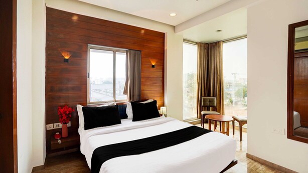 A hotel room with a large bed, wooden panel wall, two windows with curtains, a small table with chairs, and a view of the city.