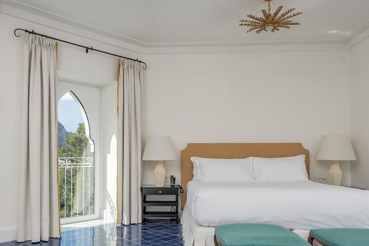 A hotel room with a bed, two bedside lamps, a wooden headboard, a small bedside table, a bench, and a view through a window.